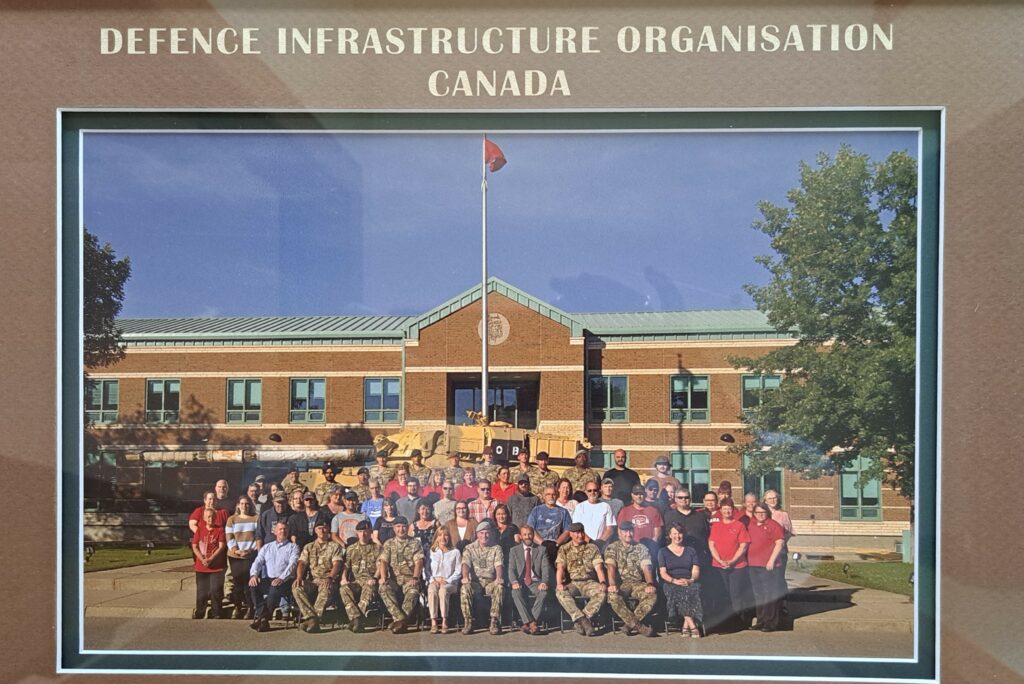 A group photo of Kate Harrison alongside various DIO and military personnel at the British Army Training Unit Suffield.