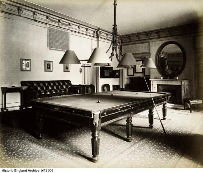 Black and white photograph of a billiard table in a room decorated in late Victorian style, with three lampshades low over the table.