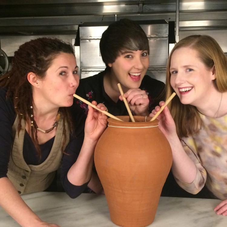 Three women gather around a large ceramic jar holding drinking straws