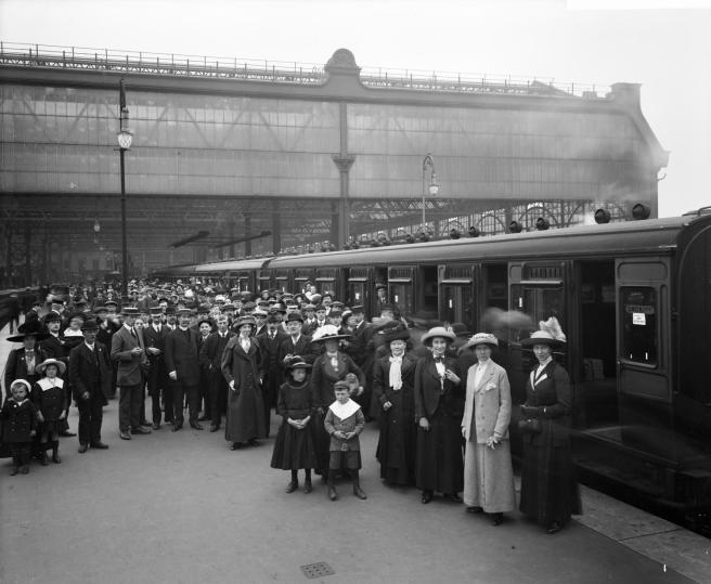 Historic B&W photograph of men, women and children posing beside a railway carriage to the left, steam rising against the roof of the station