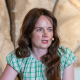Millie Horton-Insch has long, dark hair and wears a green and white top. She is speaking into a headset microphone and is sitting in front of a relief frieze. 