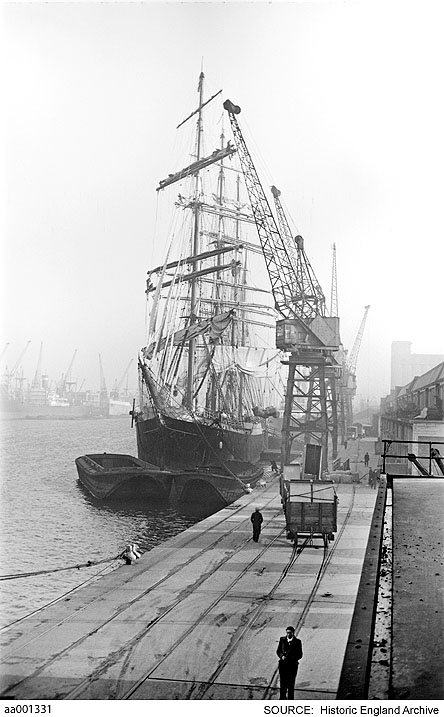B&W photo of a dockside receding to show dockside cranes. To left a tall ship whose masts reach above the dock cranes.