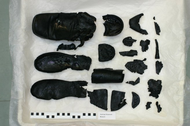 Component parts of black leather shoes, such as uppers, heels and small parts, laid out together against a white background, with a ruler at bottom left for scale.