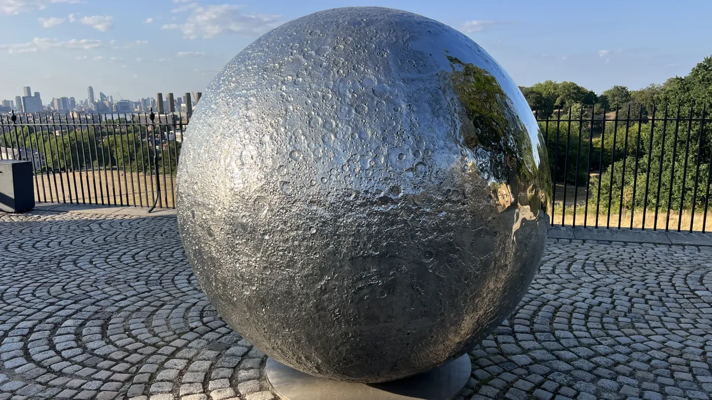 A silvery sculpture of the Moon sits in a courtyard with a cobbled floor with the view of a park in the background