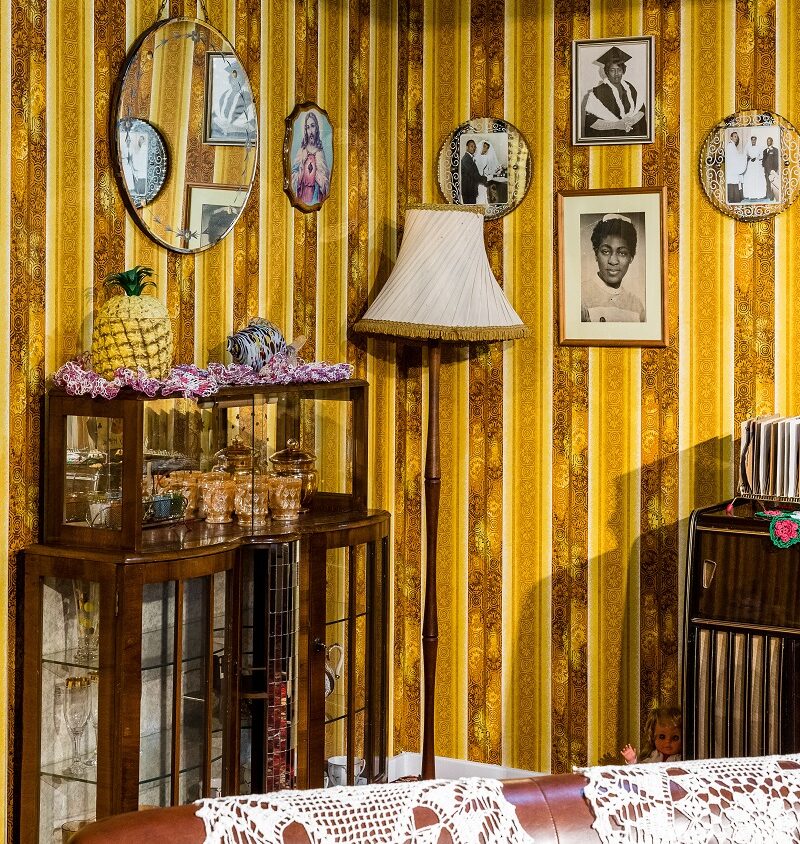 A snippet of a room display of a Caribbean British Front Room with bold yellow patterned wallpaper, framed family portraits and religious imagery, a glass-fronted cabinet with ornaments, a fringed floor lamp, and doilies on a leather sofa.