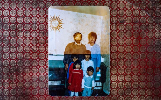 Old family photograph of two adults and four children standing in a domestic living room, looking at the camera. They are posed against patterned wallpaper with a sunburst wall clock. The photo is slightly faded and rests on a red patterned fabric background.