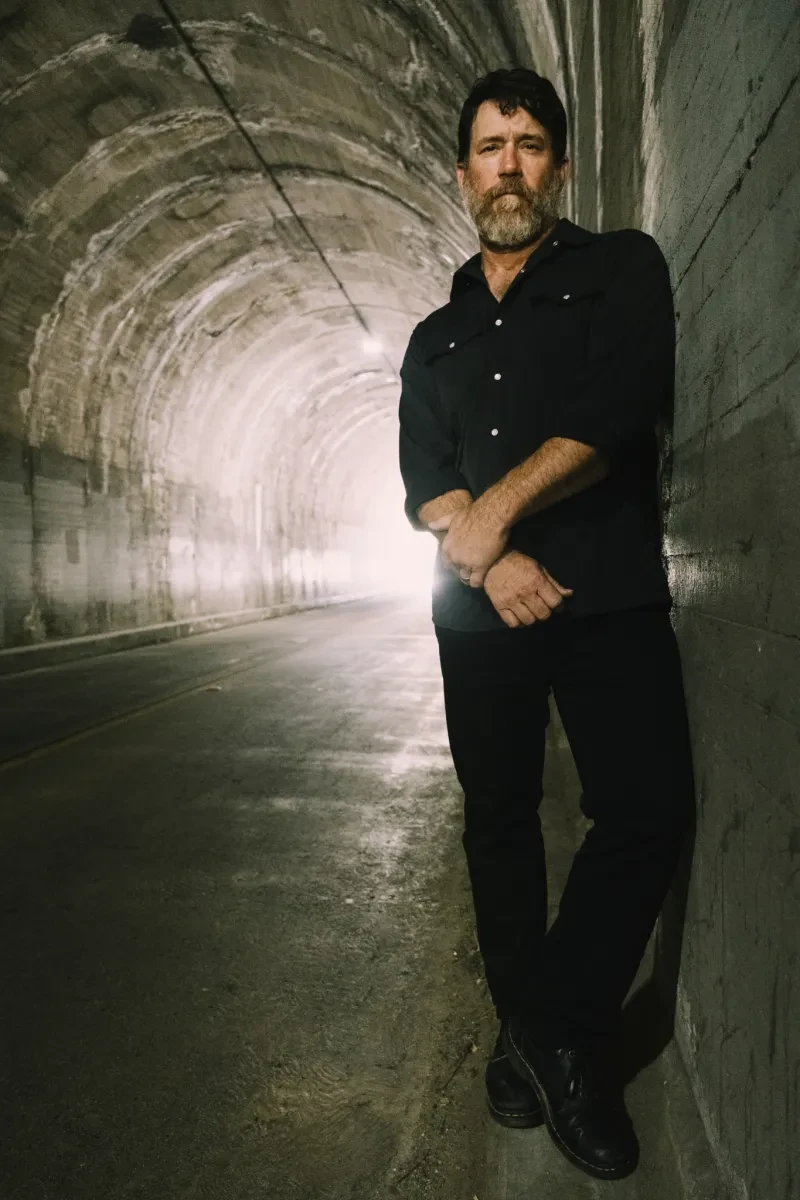 Photo of Chuck Ragan standing in a tunnel wearing black, with one hand grasping the opposite arm