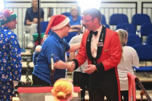 The conductor of the Royal Marines band in a red military tuxedo with black lapels wearing medals shaking the hand of chap in a Father Christmas hat in a black t-shirt. The conductor has his baton under his other arm.