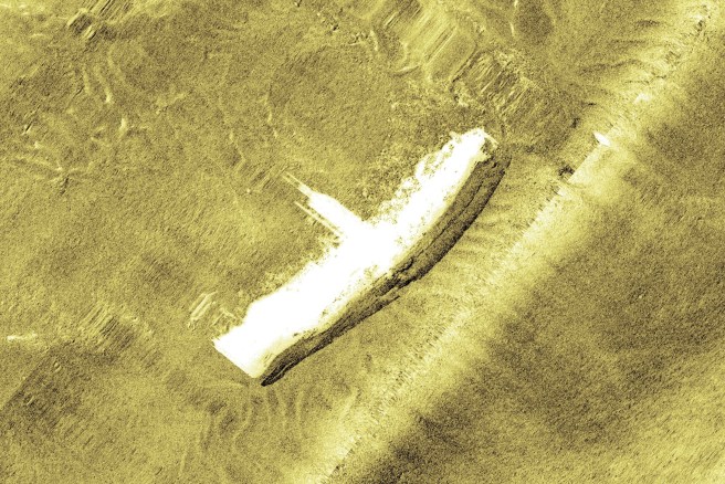 Side-scan sonar image showing vessel on its side, with its central structure clearly visible, picked out in white against an undulating textured yellowish brown background representing the seabed.