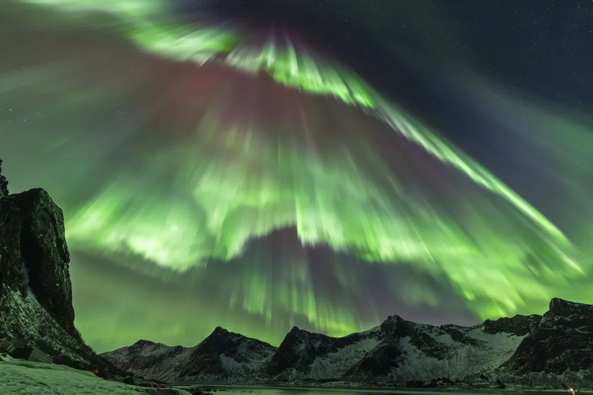 Snowy maintain range in the bottom of the image, while in the sky are several curtains of very bright and vivid green aurorae