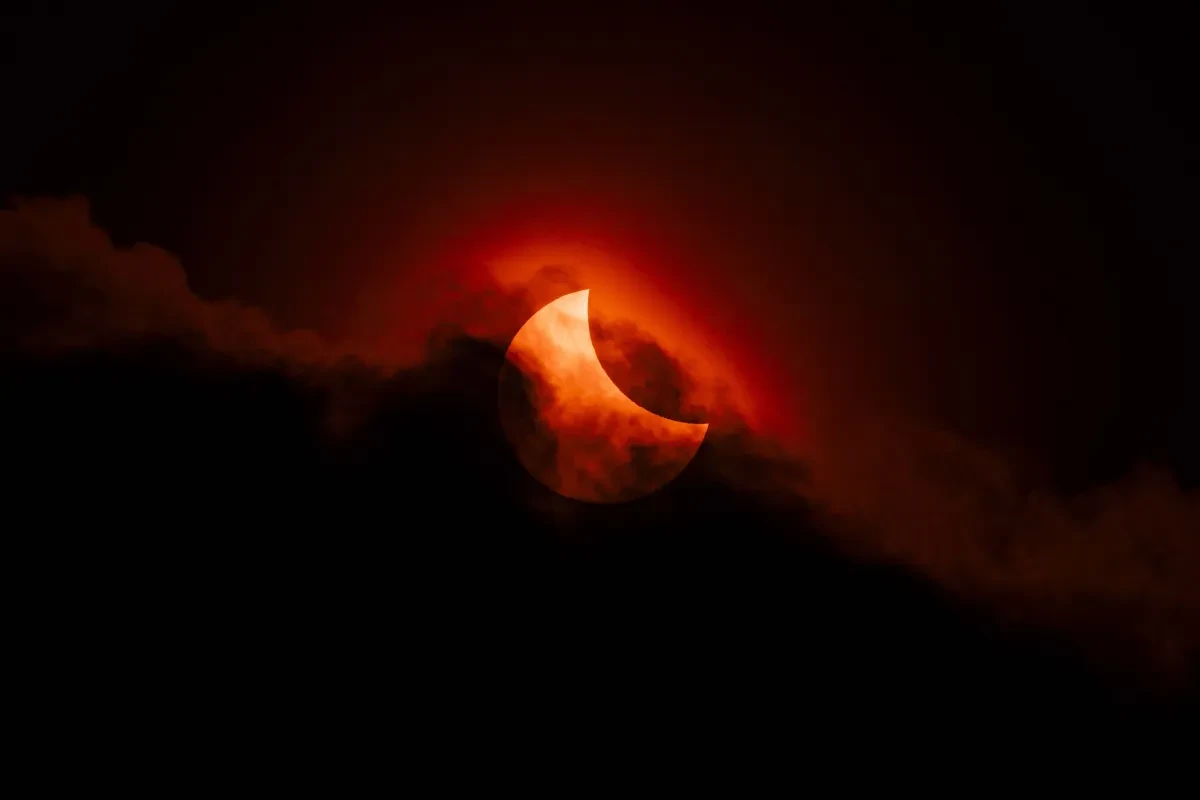 Dark image with black clouds at bottom of frame and curved dark yellow sun being eclipsed by the Moon poking out from behind wispy cloud
