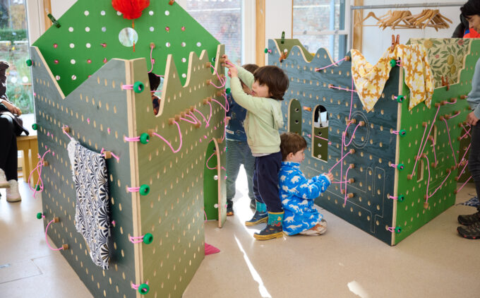 Three young children engaging with a large, green, wooden peg-board playhouse structure. Each interacting with different parts of the installation including pink elastic and bright green knob.