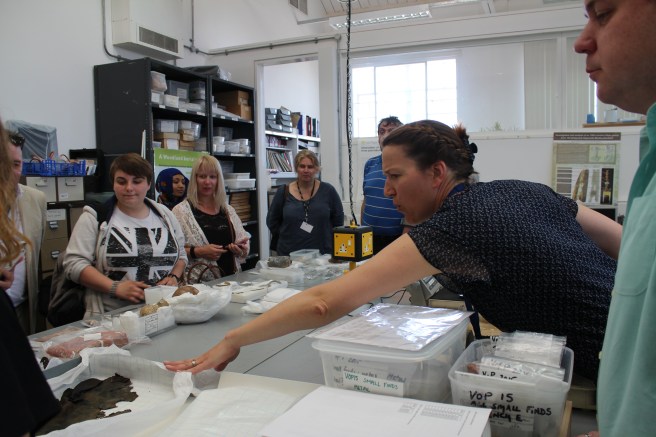 Participants standing around table in museum store listening to conservator speaking at right, with her arm stretched out across the table, which is covered with boxes and objects sitting on white conservation tissue.