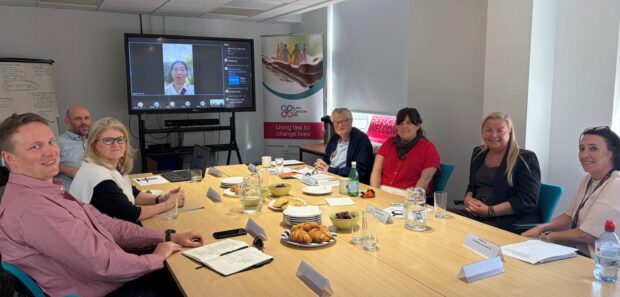 Social Security Advisory Committee (SSAC) members, Owen McCloskey and Les Allamby, seated at a table with five Northern Ireland stakeholders. A further SSAC member, Rachel Chiu, has dialled into the meeting and appears on a screen at the end of the table.