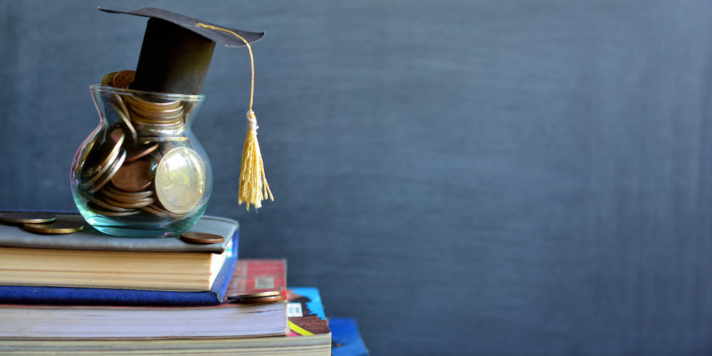 Jar of coins on a stack of books. The jar has a graduation cap on top.