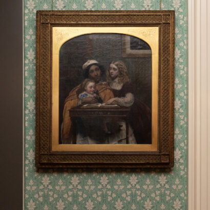 A framed painting hangs on floral-patterned wallpaper depicting a South Asian ayah—modelled by Jamaican-born Fanny Eaton—in an orange drape and white skirt, reading from a book to two white children, a toddler on her lap and an older girl leaning on the table.