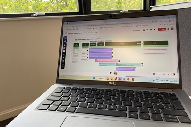 Photograph of a Dell laptop displaying a digital project management board with color-coded task cards in green, purple, and blue arranged in columns labelled with different project stages. The board includes user icons on task cards, indicating collaboration, and is viewed in a well-lit room with windows in the background.
