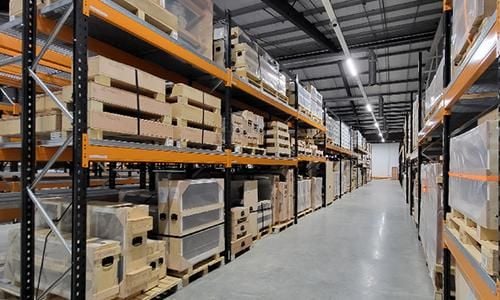  View of a large storage facility looking down an aisle with a row of shelves on either side packed with crates and storage boxes mounted on wooden pallets.