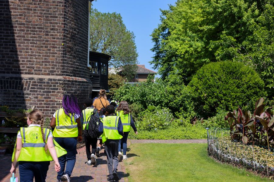 Group of young students wearing bright yellow high-visibility vests walking along a garden path beside a brick building, surrounded by trees and greenery on a sunny day.