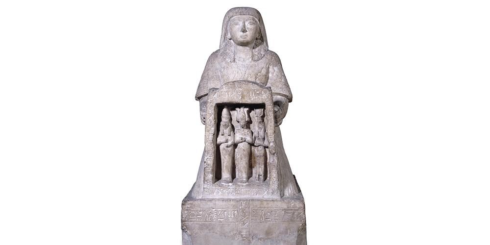A kneeling statue Panehesy who holds between his hands a naos containing the figures of Horus, Osiris and Isis and surmounted by a winged sun-disk.