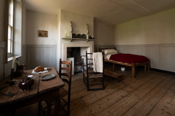 A simple almshouse room with a bed, fireplace and table with wooden floors