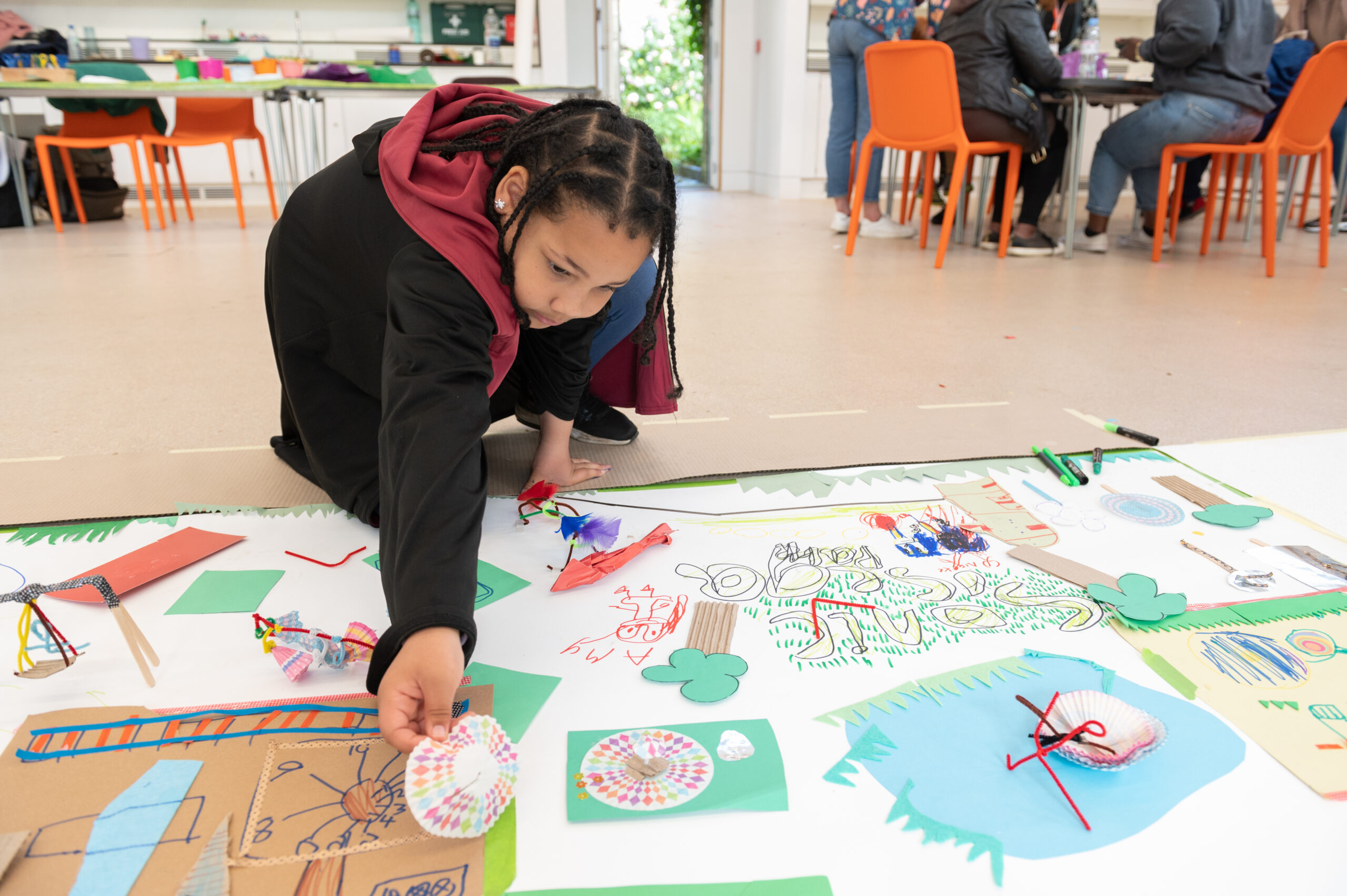 Child leaning over a large collaborative artwork on the floor, adding a colourful paper creation made from craft materials; other participants are seated at tables in the background during a creative workshop.
