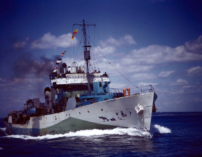 Historic colour photo of ship in starboard bow view, painted white, green and blue in dazzle camouflage, with spray at the bows as she cuts through the water under a blue sky with multiple white clouds on a fair day at sea. The paint is battered in places illustrating her hard work at sea in wartime.
