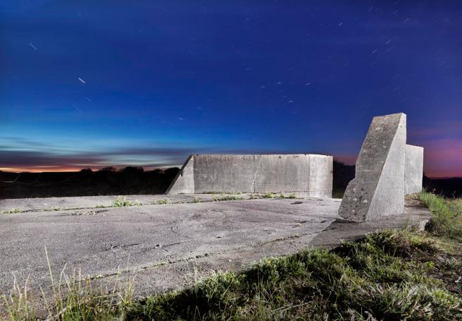 Modern colour photograph using time-lapse photography of a concrete hard standing with side features as a mock-up of the open ramp of a landing craft. It is set in grassy dunes with grass growing in the crevices between the concrete slabs.
The concrete 'landing craft' is shown at twilight with the time-lapse photography showing the movement of stars in the sky, to illustrate the time that has elapsed since it was built and had a function.