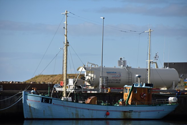 A light blue vessel is seen in broadside view berthed alongside a quay, with her small wooden cabin on deck. A white oil tanker lies behind, and behind the tanker coastal dunes are visible against a blue sky.