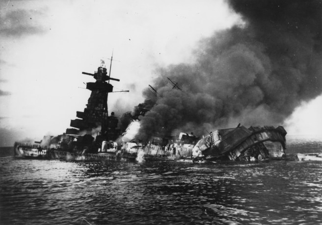 Black and white photograph of German warship on fire and sinking, with smoke billowing out of the vessel to the right.