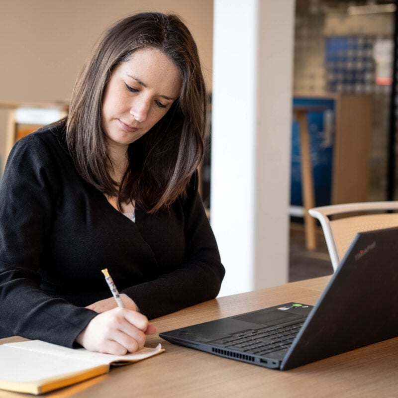 A person poised with a pen taking notes in a notebook. They are in deep thought sitting at a desk in what appears to be an office with a laptop open in front of her.