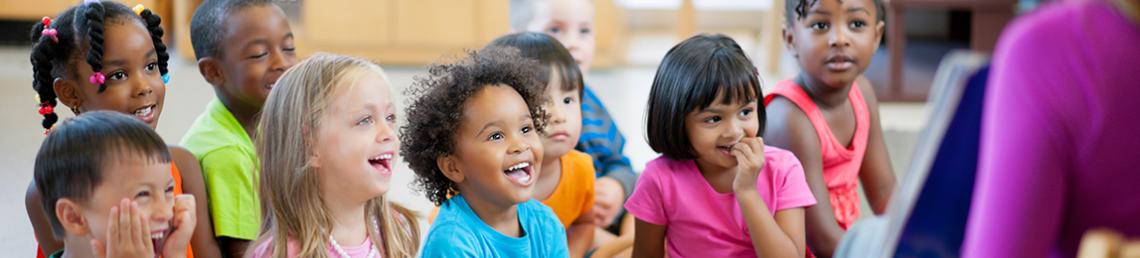 A mixed group of young children looking very happy while listening to someone reading a book 