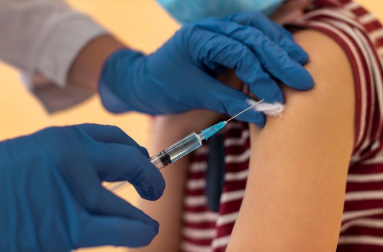A medical professional with blue gloves on giving a young person a vaccine in their arm, the person has a stripy red top on. 