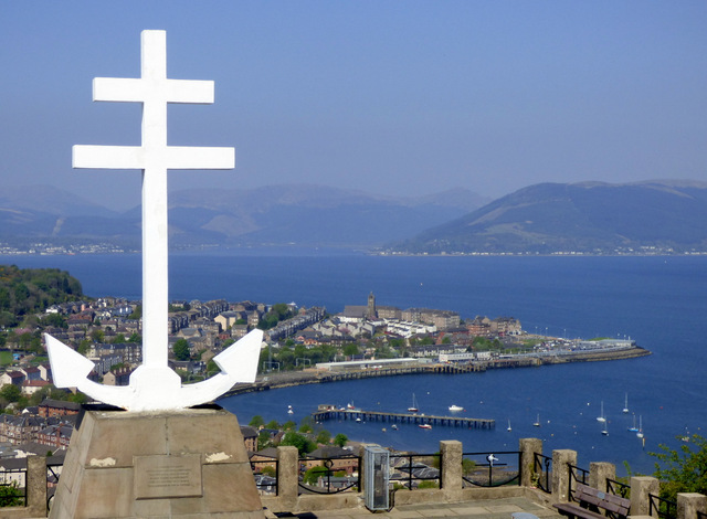 Modern colour photograph of naval memorial in white to left of image, overlooking a harbour with blue sea, blue hills and blue sky in the background. 