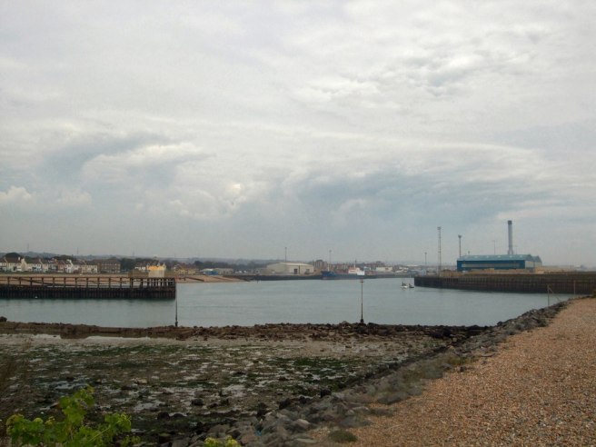 Modern colour photo taken from the viewpoint of the shingle bank in the foreground and to right which encloses the canal from the sea, looking to both arms of the canal east and west, under a heavy greyish sky. 