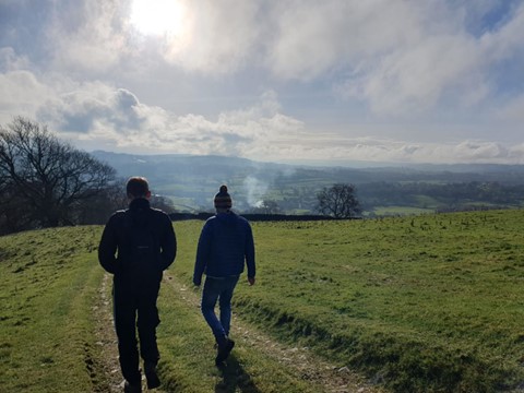 Two silhouetted figures walk downhill on a footpath within a green field. Farmland can be seen on the distant horizon for miles. Clouds hang overhead.