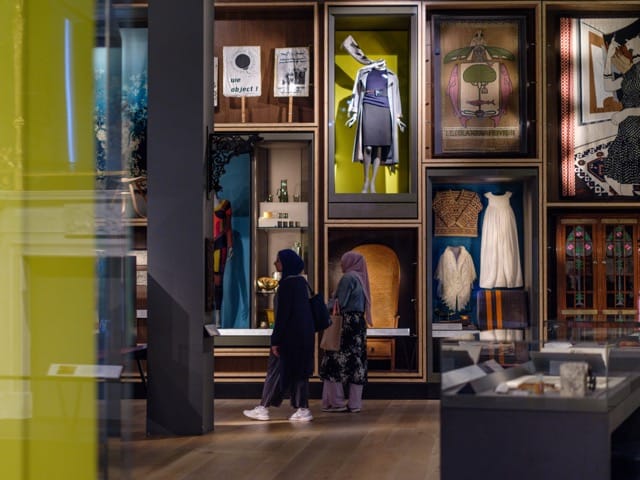 Two women view diverse exhibits in a museum, with fashion and art pieces displayed in illuminated cases against a colorful background.