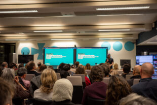 Rows of seated audience members listening to a talk being delivered in front of a presentation screen