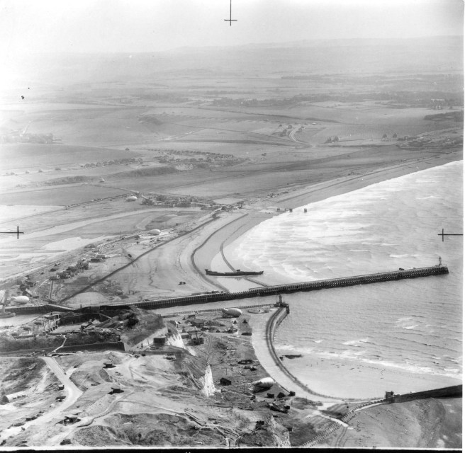 Aerial photograph of Newhaven to foreground, pier to centre ground, wreck across beach behind the pier, and the rural coastline with patchwork fields and beach stretching away beyond. 