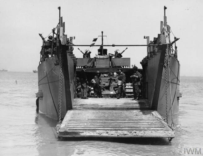 Historic B&W photo of Landing Craft Tank ramp down in shallow water, with soldiers and tank preparing to exit. There are guns visible at the sides and on top of the vessel's bridge in the background to provide covering fire. 