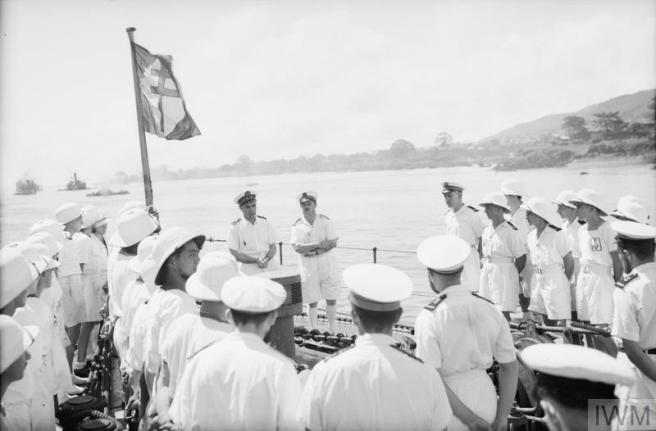 Historic B&W photograph of seamen in white tropical uniforms gathering to listen to a speaker on board ship under the Free French flag towards top left. Other ships are visible in the distance to left beyond the flag. 