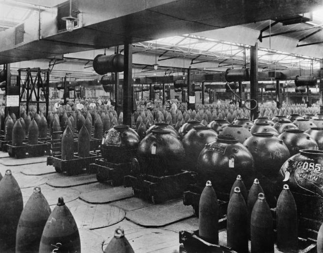 Historic B&W photograph of long rows and rows of shells and mines of different types loaded on trolleys in a large factory. 