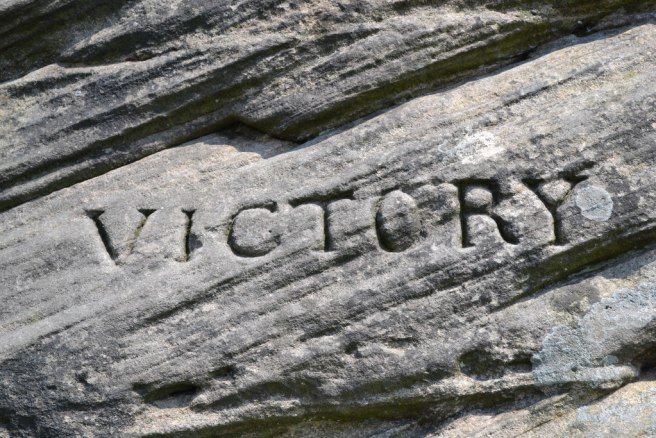 Modern colour photo of the word VICTORY incised on a rock and still legible despite erosion. The rock formations run from bottom left to top right of the image, are weathered in places, and have light coloured spots of lichen. 
