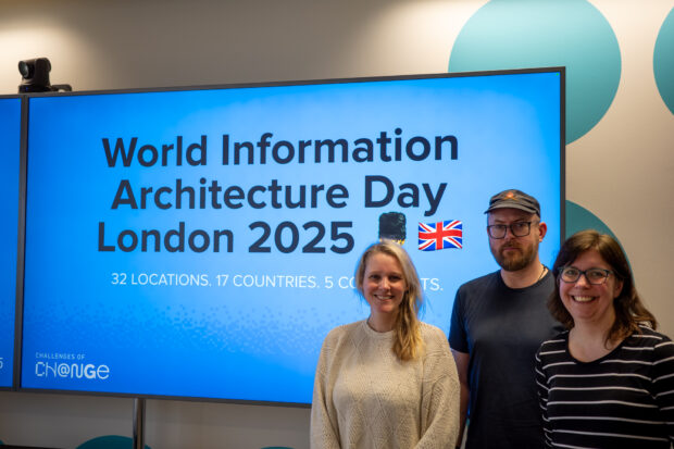 Three information architects standing in front of a large screen displaying a presentation slide which reads: “World Information Architecture Day London 2025. 32 locations, 17 countries, 5 continents.”