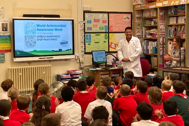 Image of a male in a white lab coat standing in a classroom of children who are sitting on the floor in front of him. There is a screen at the front which says, "World Antimicrobial Awareness Weekn 18-24 November, Spread Awareness Stop Resistance"