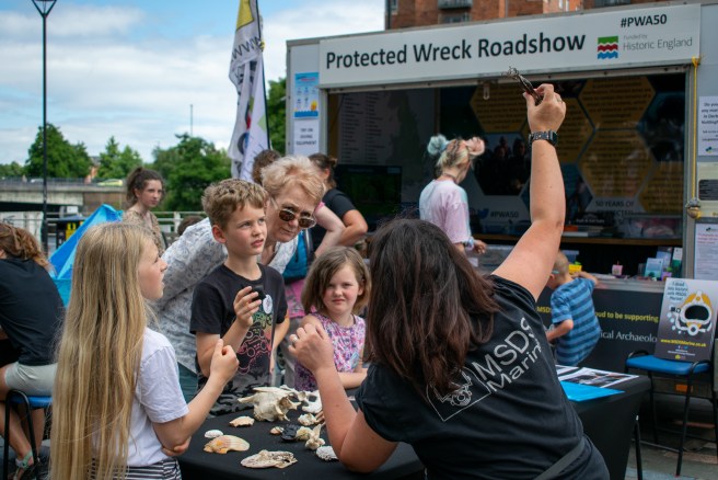 Visitors of all ages explore shells, bones and artefacts from the sea with a helper from MSDS Marine: a Protected Wreck Roadshow banner is in the background 