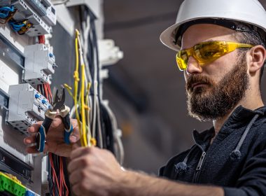 A Male Electrician Works In A Switchboard With An Electrical Connecting Cable.