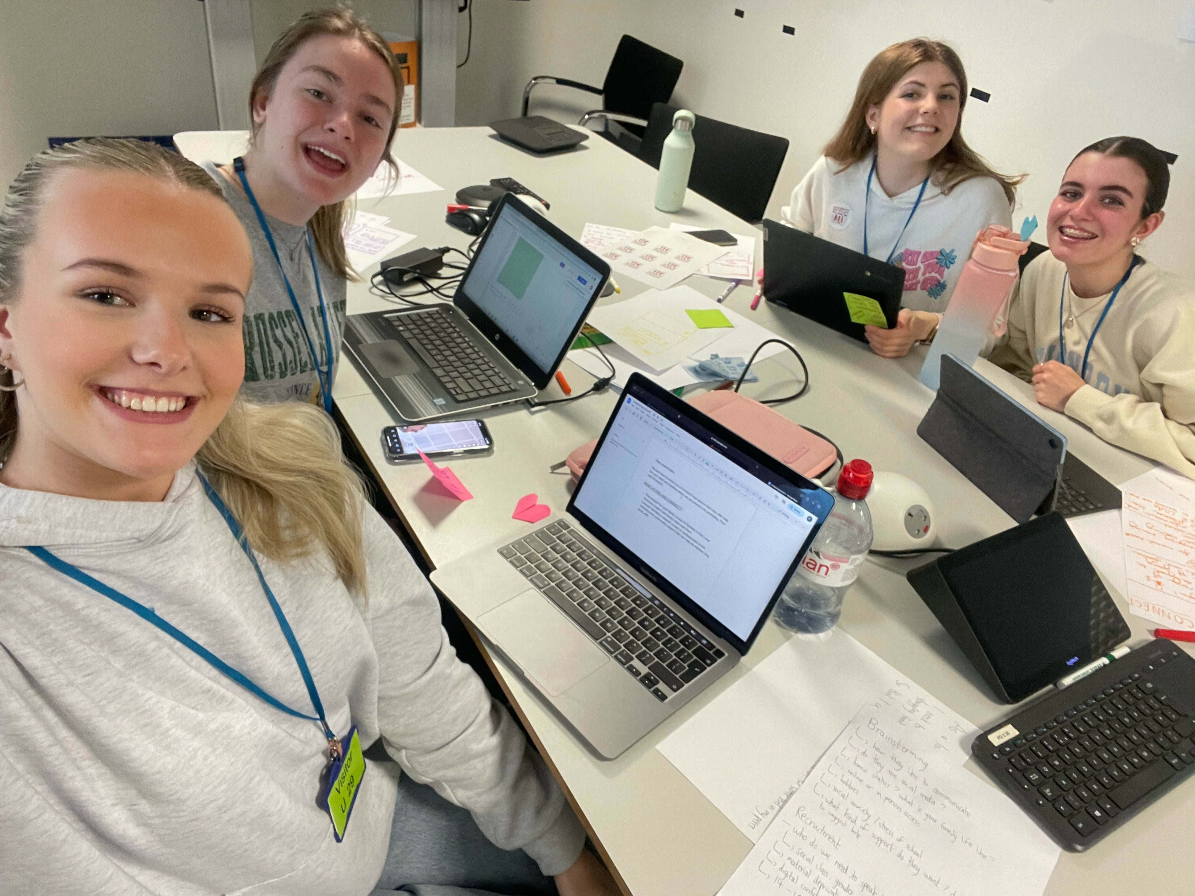 Group of four teenagers gathered around a conference table with laptops and office paraphernalia