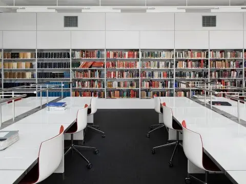 IWM’s reading room. The room is full of long white desks with swivel chairs. Running along one wall are shelves full of reference books for consultation.