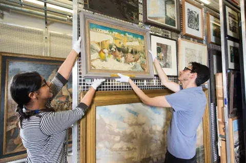 Two IWM staff reaching for a painting in the art store
